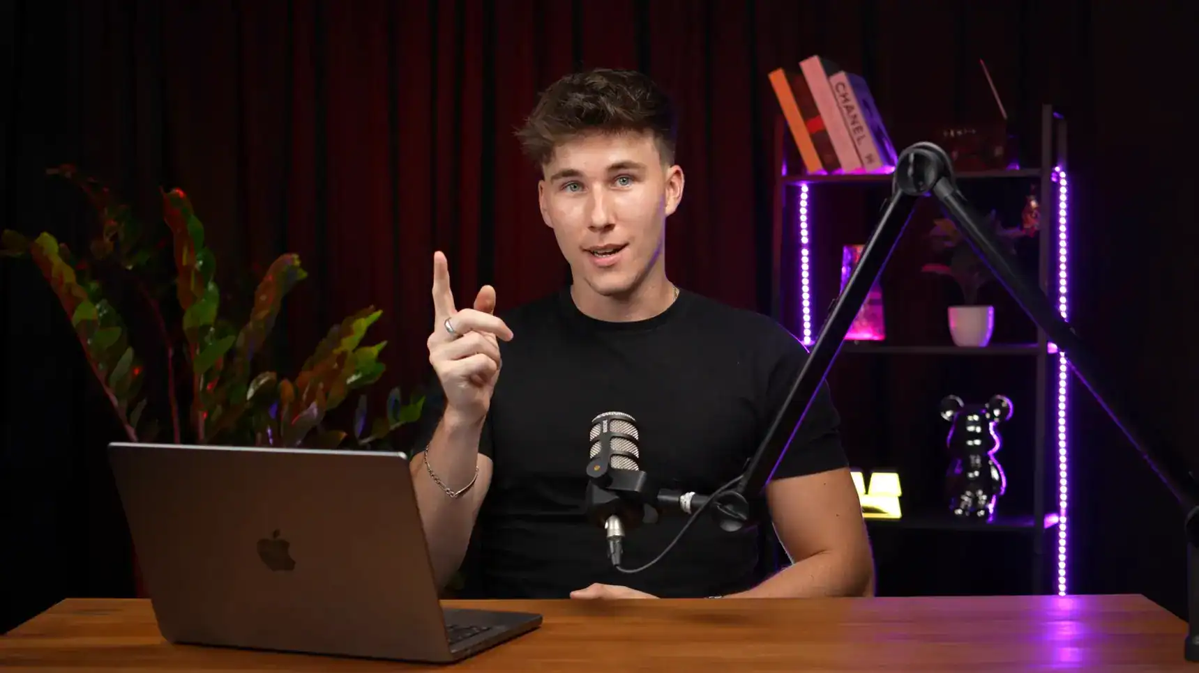 Brightly lit podcast studio featuring a young male host, microphone, MacBook, and stylish shelving with decor and books.