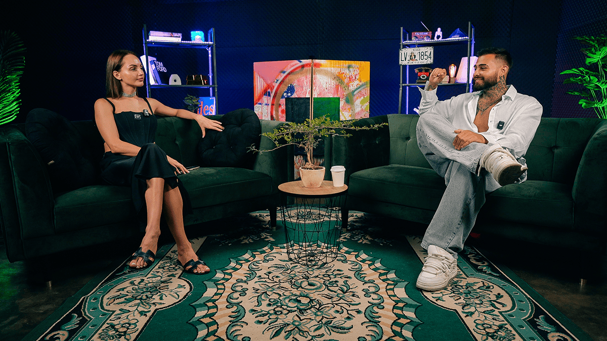Relaxed studio interview setting with two people on plush green sofas.